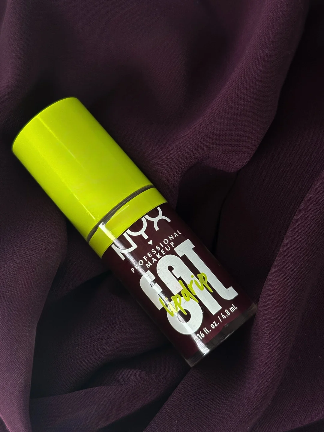 Single NYX Professional Makeup Fat Oil Lip Drip bottle with chartreuse cap on deep plum fabric.